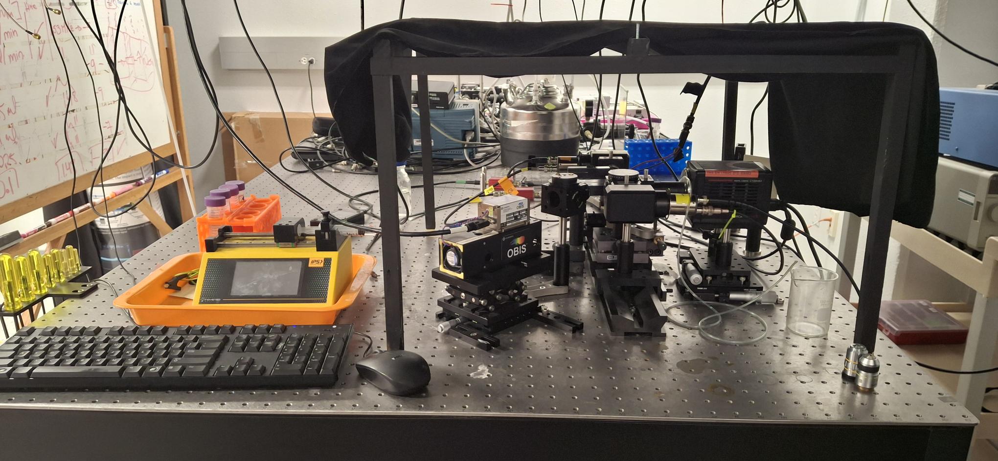 Optical set up of the TRFFC instrument on the optical table with the exposed optical components for laser alignment presented. Including the syringe pump to deliver the sample to the microfluidic chip for measurement and acoustic focusing.