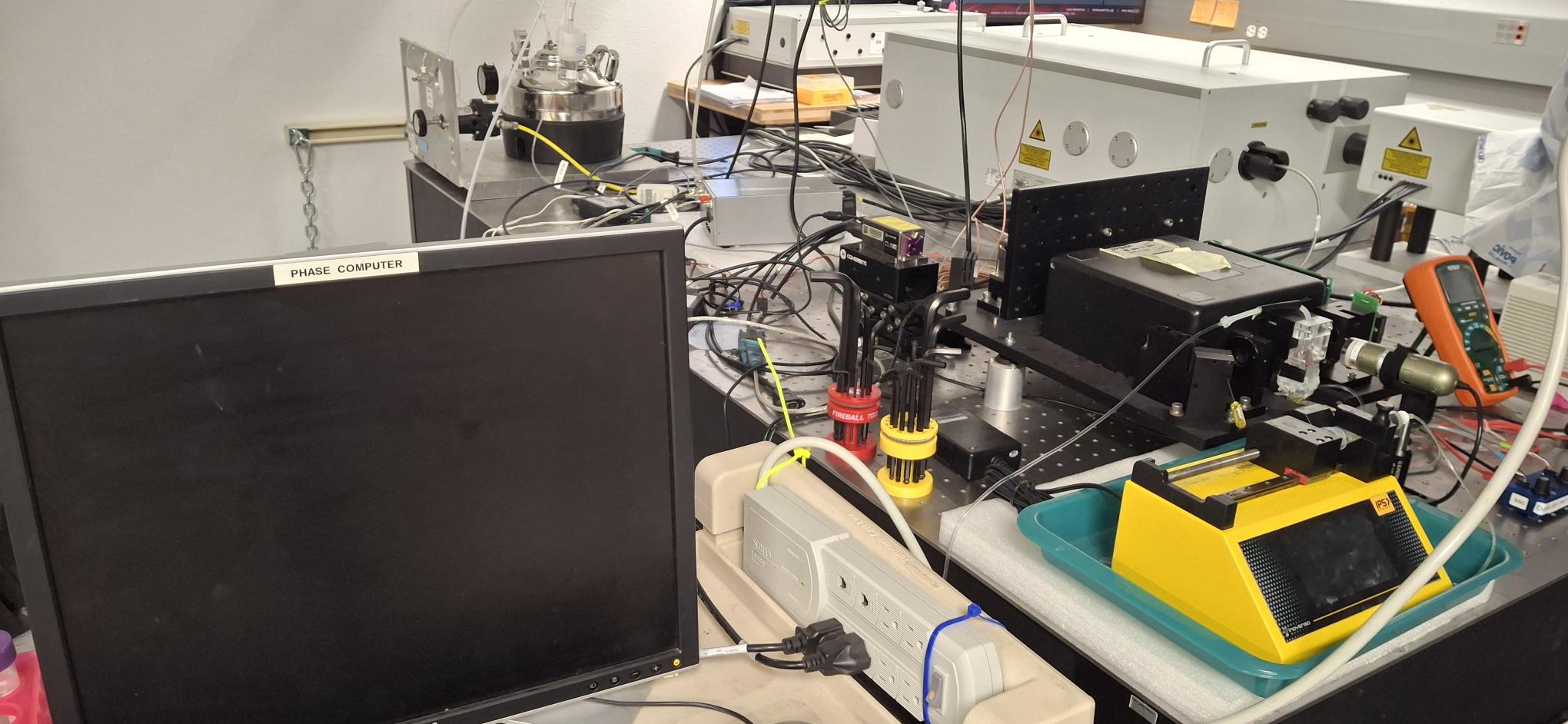 The computer screen next to the TRFC instrument on the optical table. The syting pump used to deliver sample to the instrument is also in the image. The TRFC instrument is on an elevated platform and most of it is covered by a black box. However, there are two mirrors are visible, as is the flow cell with a camera in front of the flow cell.